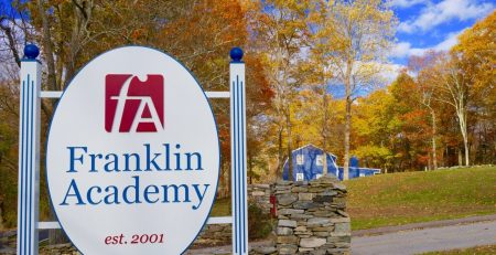 The entrance sign at Franklin Academy, with the school's logo, an "F" and "A" inside a red square, and the words "Franklin Academy" in blue. In the background are the yellows and oranges of New England fall foliage.