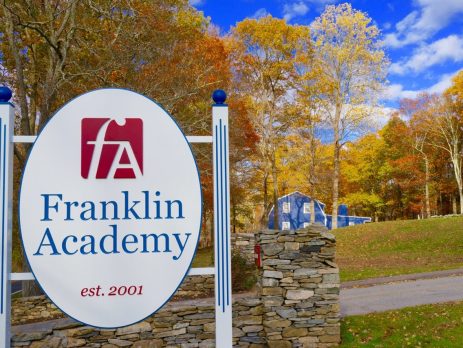 The entrance sign at Franklin Academy, with the school's logo, an "F" and "A" inside a red square, and the words "Franklin Academy" in blue. In the background are the yellows and oranges of New England fall foliage.