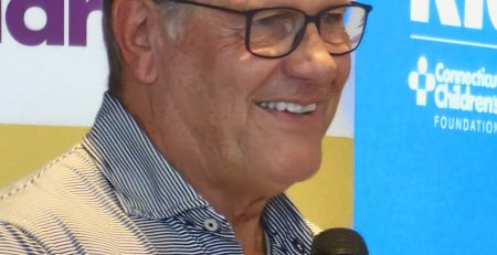 UConn women's basketball coach Geno Auriemma, a white man wearing black glasses and a plain gray button-down shirt, holding a microphone. In the background are the two logos of Connecticut Children's Medical Hospital and Geno For The Kids.