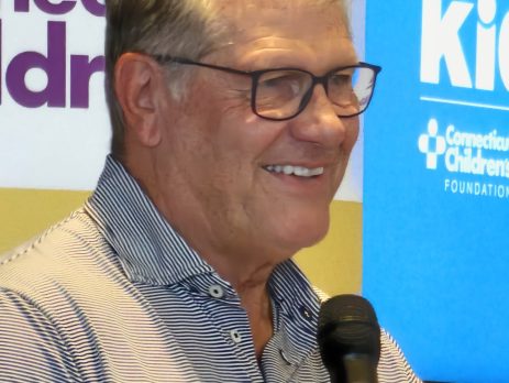 UConn women's basketball coach Geno Auriemma, a white man wearing black glasses and a plain gray button-down shirt, holding a microphone. In the background are the two logos of Connecticut Children's Medical Hospital and Geno For The Kids.