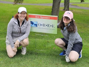 Mary and Kelly Coons smiling and kneeling at the OREE Ace Sponsor sign at the Watkinson Golf Classic 2024.