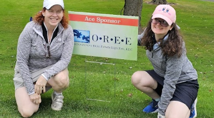 Mary and Kelly Coons smiling and kneeling at the OREE Ace Sponsor sign at the Watkinson Golf Classic 2024.