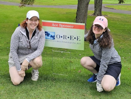 Mary and Kelly Coons smiling and kneeling at the OREE Ace Sponsor sign at the Watkinson Golf Classic 2024.