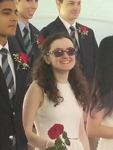 Kelly Coons in a white dress at her high school graduation, holding a red rose and smiling.