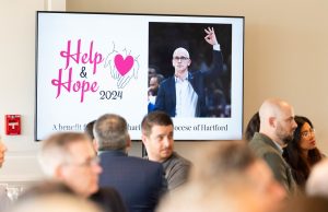 A poster in the background, with the Help & Hope Breakfast 2024 logo, and a picture of UConn Huskies head coach Dan Hurley celebrating a play with the OK sign.