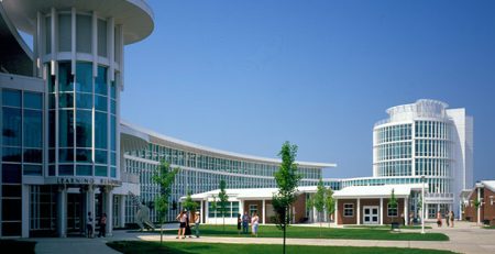 The campus of Connecticut State Community College (previously known as Manchester Community College) on a clear, blue day. The photo is taken from a 3/4 angle, with elegant, sleek, white, many-windowed buildings on the left side. The landscape is dotted with trees.