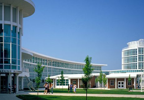 The campus of Connecticut State Community College (previously known as Manchester Community College) on a clear, blue day. The photo is taken from a 3/4 angle, with elegant, sleek, white, many-windowed buildings on the left side. The landscape is dotted with trees.